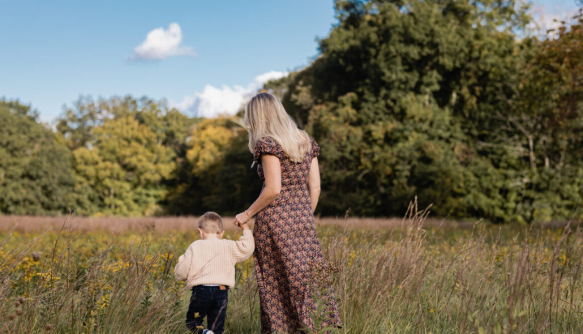 woman in floral print dress holds blonde son in oatmeal colored sweater as they walk hand in hand through a field of wild flowers during golden hour in autumn