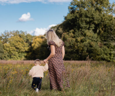 woman in floral print dress holds blonde son in oatmeal colored sweater as they walk hand in hand through a field of wild flowers during golden hour in autumn