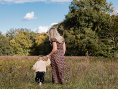 woman in floral print dress holds blonde son in oatmeal colored sweater as they walk hand in hand through a field of wild flowers during golden hour in autumn