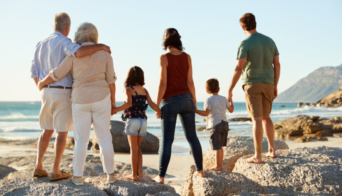 Three generation white family on a beach stand holding hands, admiring view, full length, back view