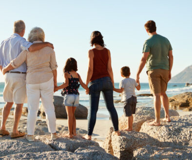 Three generation white family on a beach stand holding hands, admiring view, full length, back view