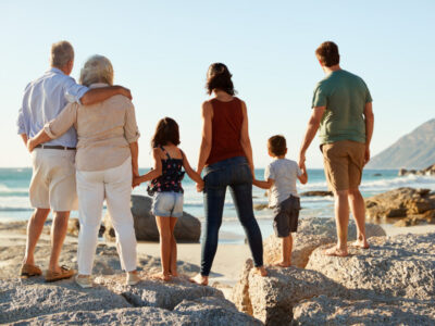 Three generation white family on a beach stand holding hands, admiring view, full length, back view